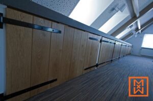 Op maat gemaakte, licht eikenhouten opbergkasten met zwart metalen schuurdeurbeslag sieren een schuin aflopende zolderkamer. De kasten sluiten naadloos aan op het moderne design van de kamer en bieden stijlvolle opbergoplossingen.