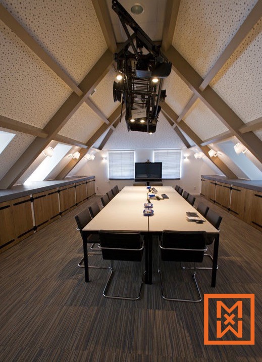 Moderne, minimalistische vergaderruimte met een lange witte tafel en zwarte stoelen. Aan het hoge, schuine plafond hangt een unieke lamp in industriële stijl. De ruimte is voorzien van houten lambrisering en een neutraal kleurenpalet.