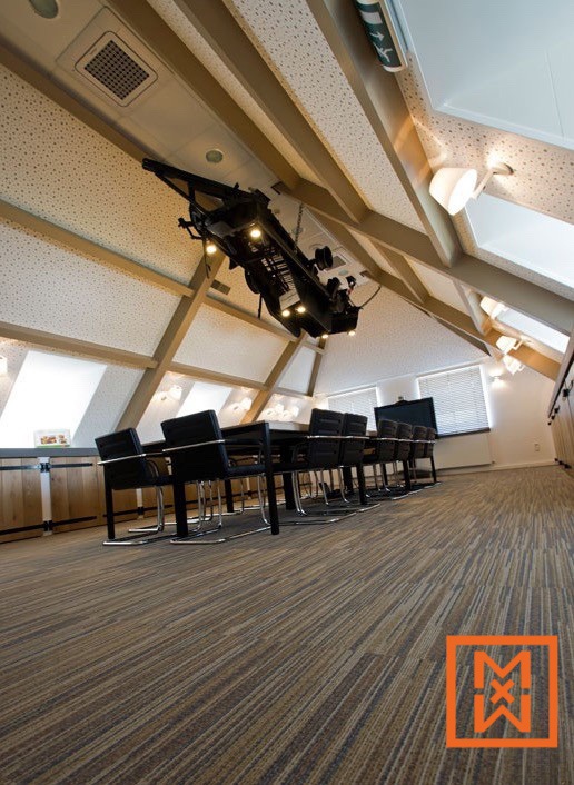 Een moderne vergaderzaal met een uniek design. De kamer heeft een lange conferentietafel, zwarte stoelen en een opvallend plafond met een projector. De vloer is bedekt met bruin tapijt. Een perfecte inspiratie voor modern office design.