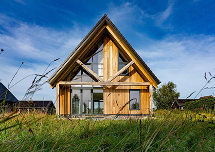 Houten huis met een driehoekige gevel en grote ramen, omgeven door groen gras en een blauwe lucht. Modern design in een landelijke omgeving.