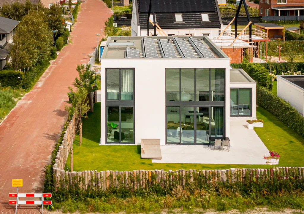 Moderne, witte woning met zonnepanelen en grote ramen, omgeven door een groene tuin en een houten hek. Een rustige woonwijk met een oranje straat.