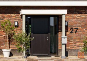 Moderne voordeur van een bakstenen huis met huisnummer 27. De deur is zwart met glazen panelen, omlijst door grijze pilaren en een witte bovenlijst. Kleine boom in pot links.
