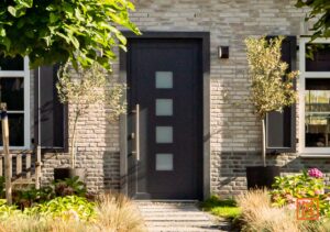 Moderne voordeur met vierkante ramen in een stenen huis. Groene planten en bomen omlijsten de ingang, met een pad van stenen platen dat naar de deur leidt.