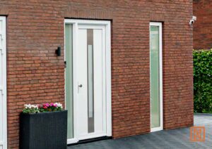 Witte voordeur met verticaal raam in bakstenen muur, naast smal raam en donkere bloembak met bloemen. Beveiligingscamera zichtbaar. Moderne woning in Nederland.