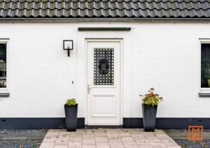 Witte bakstenen gevel met een witte voordeur met sierlijk rooster. Twee zwarte plantenbakken flankeren de ingang. Zwarte dakpannen en een moderne buitenlamp completeren het beeld.