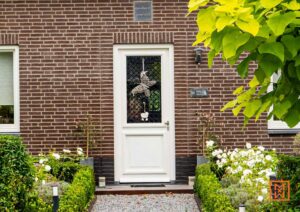 Een uitnodigende voordeur in een bakstenen huis met een witte deur en decoratieve elementen. De tuin is weelderig groen met witte bloemen. Woerdshof 10, anno 2018 is te lezen boven de deur.