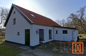 Charmante gerenoveerde boerderij met een rood pannendak en witgekalkte muren, gelegen in een rustige, landelijke omgeving. De woning heeft een donkergroene deur en diverse ramen met donkere kozijnen, die het vernieuwde ontwerp benadrukken.