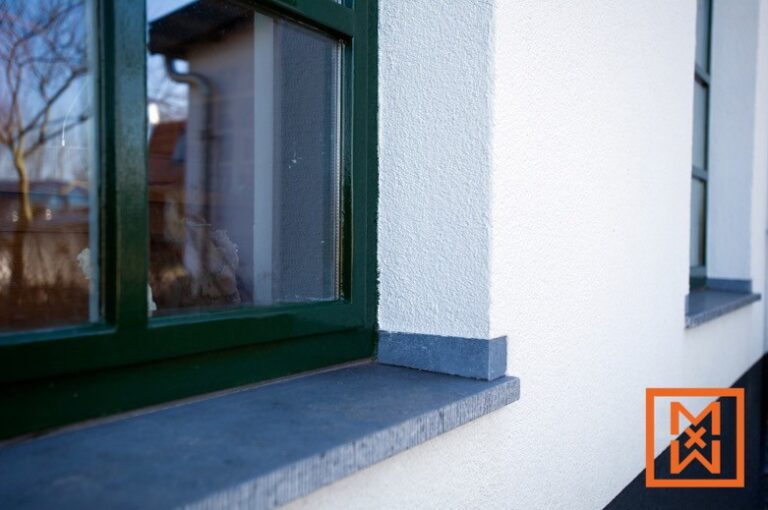 Close-up van de buitenkant van een modern huis, met een donkergroen raam met een vensterbank van leisteen. De strakwitte stucmuur zorgt voor een strak contrast. De foto benadrukt de minutieuze details aan de buitenkant.