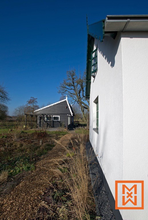 Een schilderachtig, donkergrijs huis in Nederlandse stijl met witte randen staat naast een witgekalkt gebouw op een zonnige dag. De scène omvat een tuin met gedroogd gras en mulch, wat een landelijke omgeving suggereert.