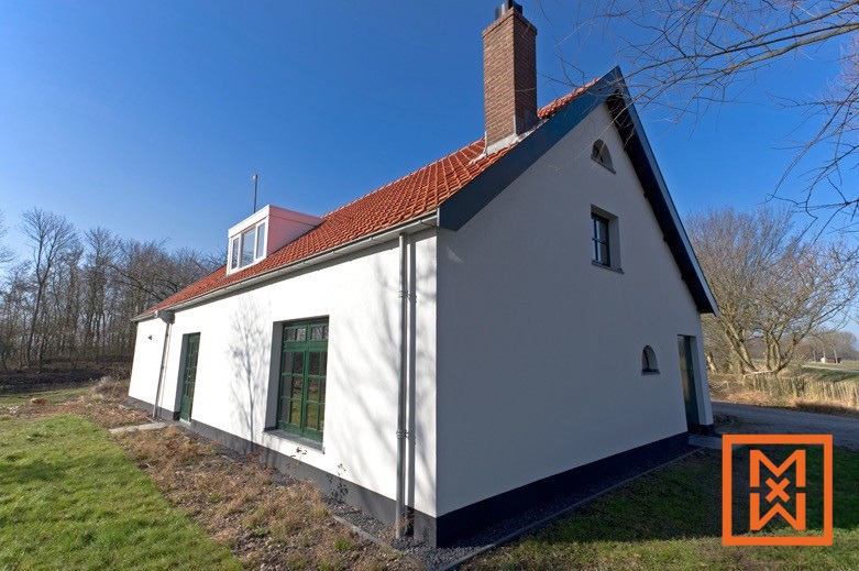 Onlangs gerenoveerde witte boerderij met een rood pannendak, donkergroene ramen en een dakkapel. Het huis ligt op een grasveld, met bomen en een helderblauwe lucht op de achtergrond.